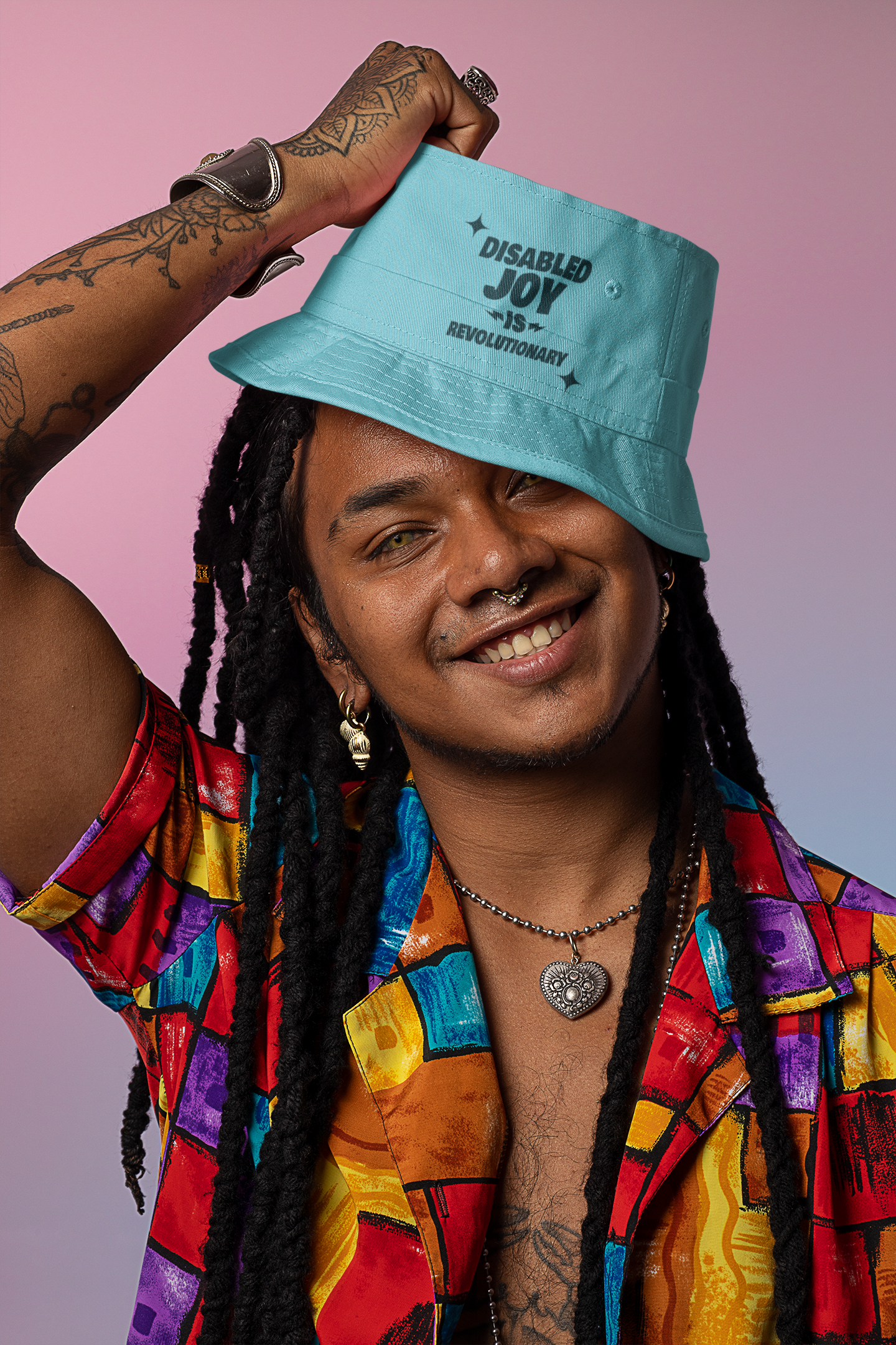 A person in a colorful shirt and a teal bucket hat with "Disabled Joy is Revolutionary" written on it, smiles and lifts the hat slightly. They have long braids, tattoos, and various jewelry. The background is pink and purple.