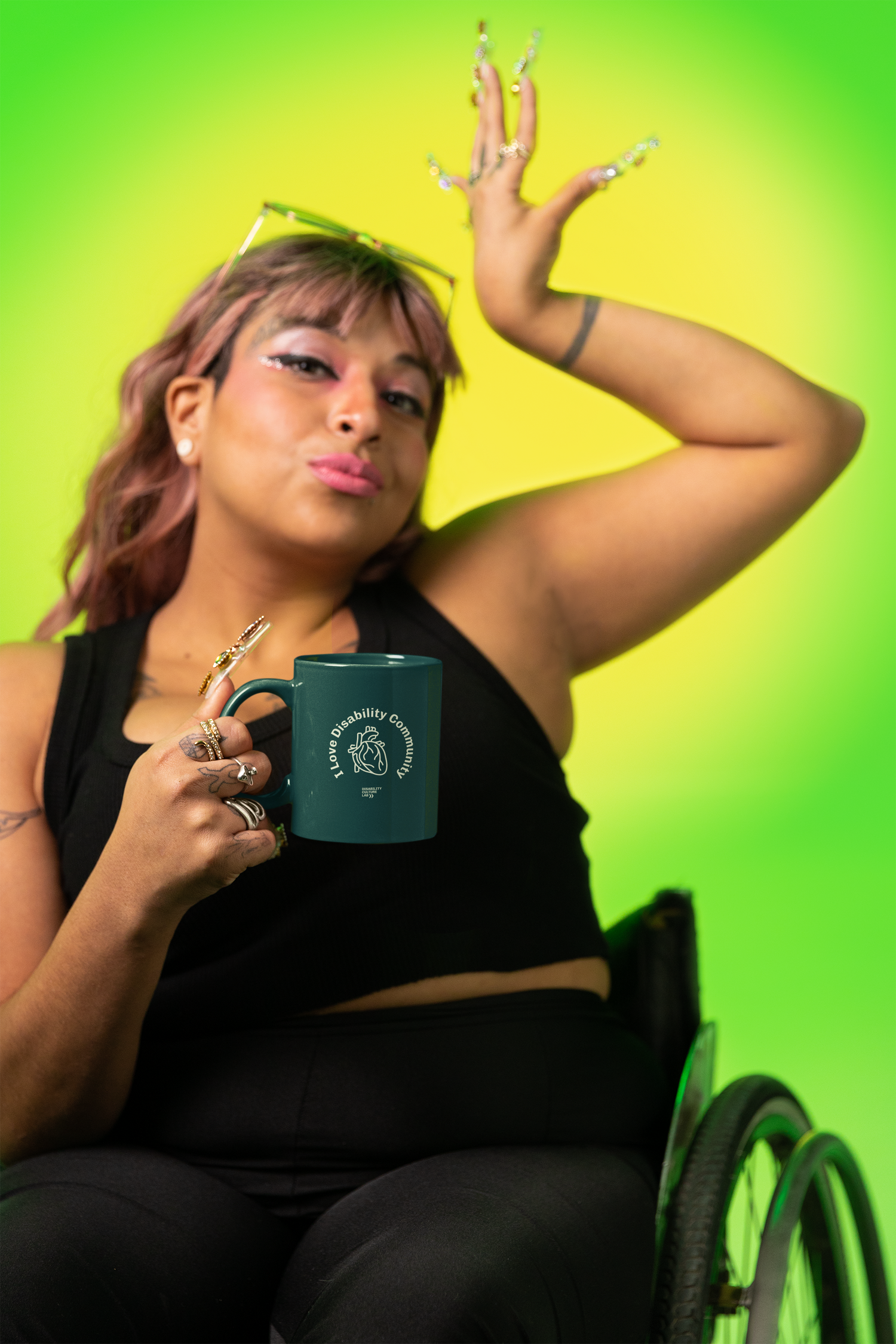 A person in a wheelchair holds a mug with the words "Do The Accessibility Consent Thing" set against a vibrant green background. They pose confidently, wearing a black outfit and stylish sunglasses on their head.