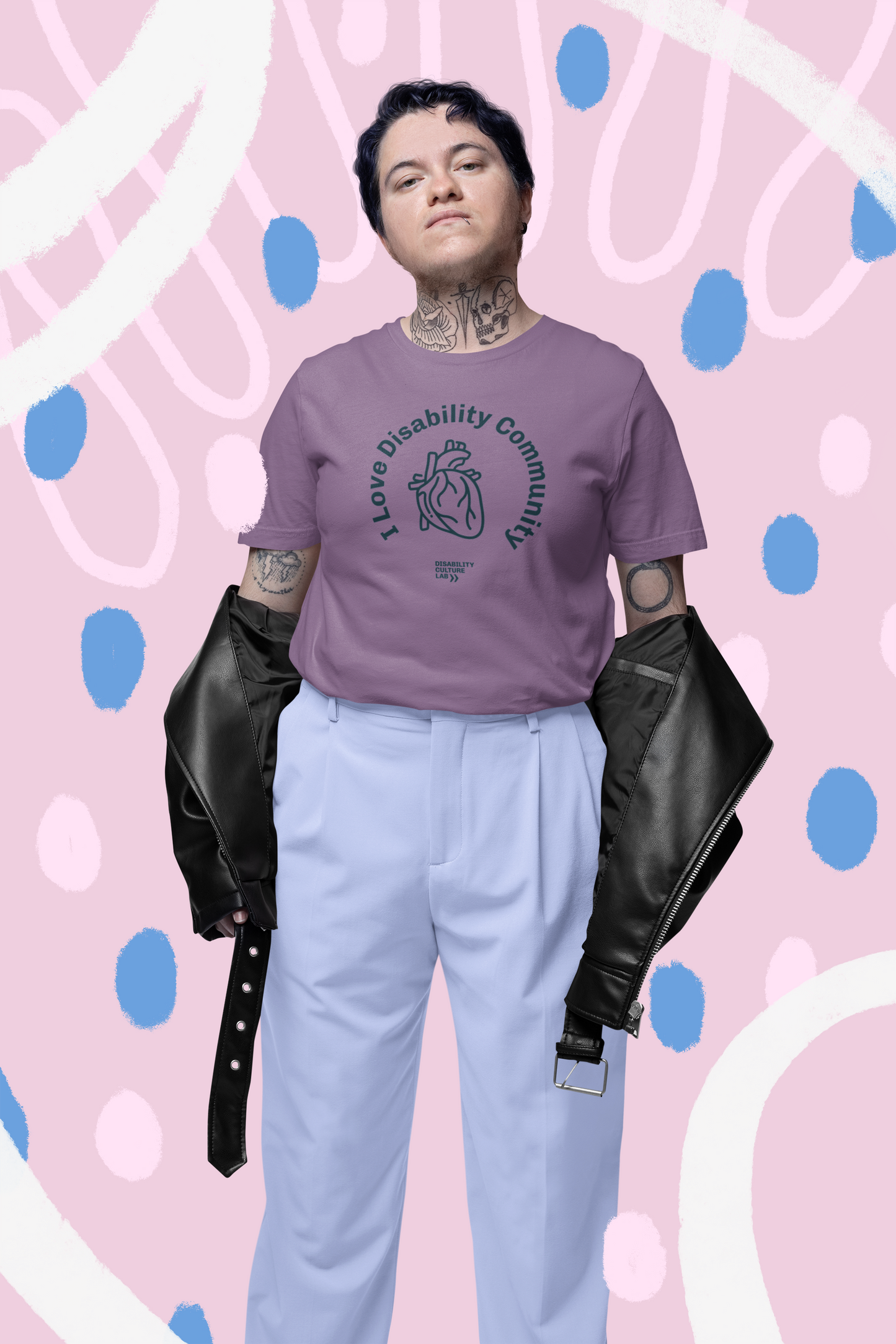 A tattooed individual stands confidently against a pastel, abstract background. They are wearing the "I Heart Disability Community" T-shirt from Disability Culture Lab, draped with a black jacket over their arms, and paired with light blue pants.