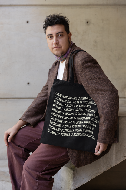 A person with short, curly hair sits on a step against a concrete wall, wearing a brown blazer and purple pants. They hold the "Disability Culture Is" tote bag by Disability Culture Lab, made of 100% cotton, featuring white text about disability justice by Jennifer White-Johnson and its connection to various social issues.