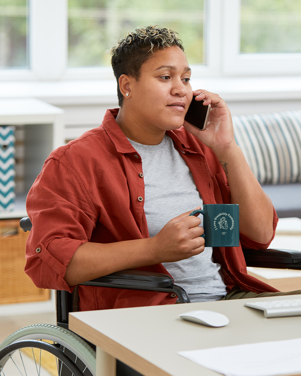 A person in a wheelchair, embodying Disability Culture from the Disability Culture Lab, talks on the phone while holding an "I Heart Disability" Ceramic Mug. They are dressed in a red shirt layered over a gray t-shirt. Amidst paperwork on the desk is a computer mouse with a design style similar to that of Jennifer White-Johnson.