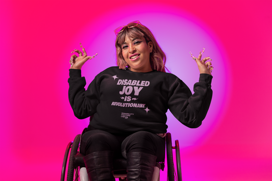 A person in a wheelchair beams with pride, wearing the Disability Joy Sweatshirt by Disability Culture Lab. They show off long, decorated nails and sport pink sunglasses perched on their head against a pink backdrop with bright circular light.