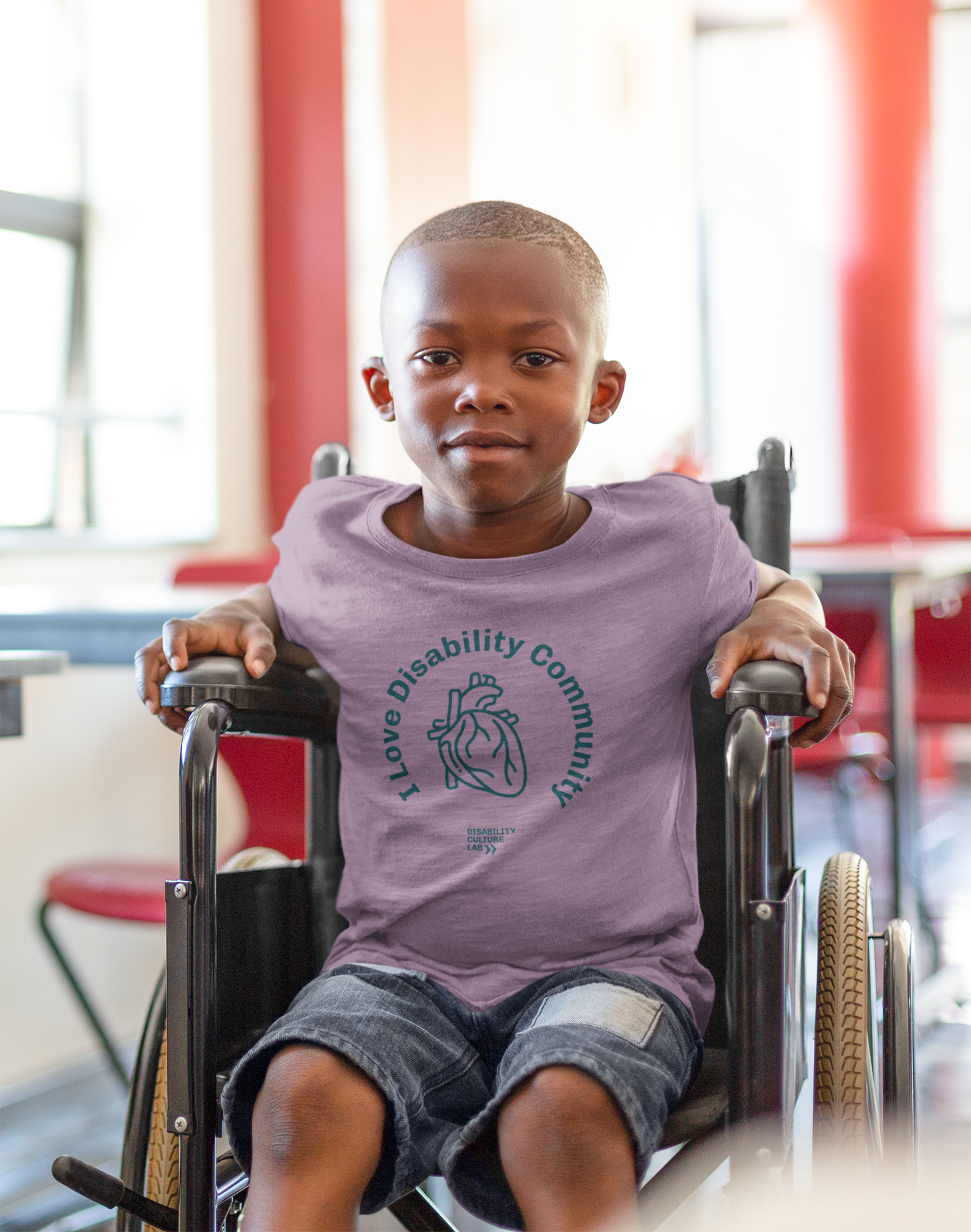 A young child sits in a wheelchair, wearing the I Heart Disability Community Kids Tee by Disability Culture Lab, a limited-edition piece that highlights the creativity of a talented disabled artist. The red and white walls and large windows in the background indicate an indoor location.