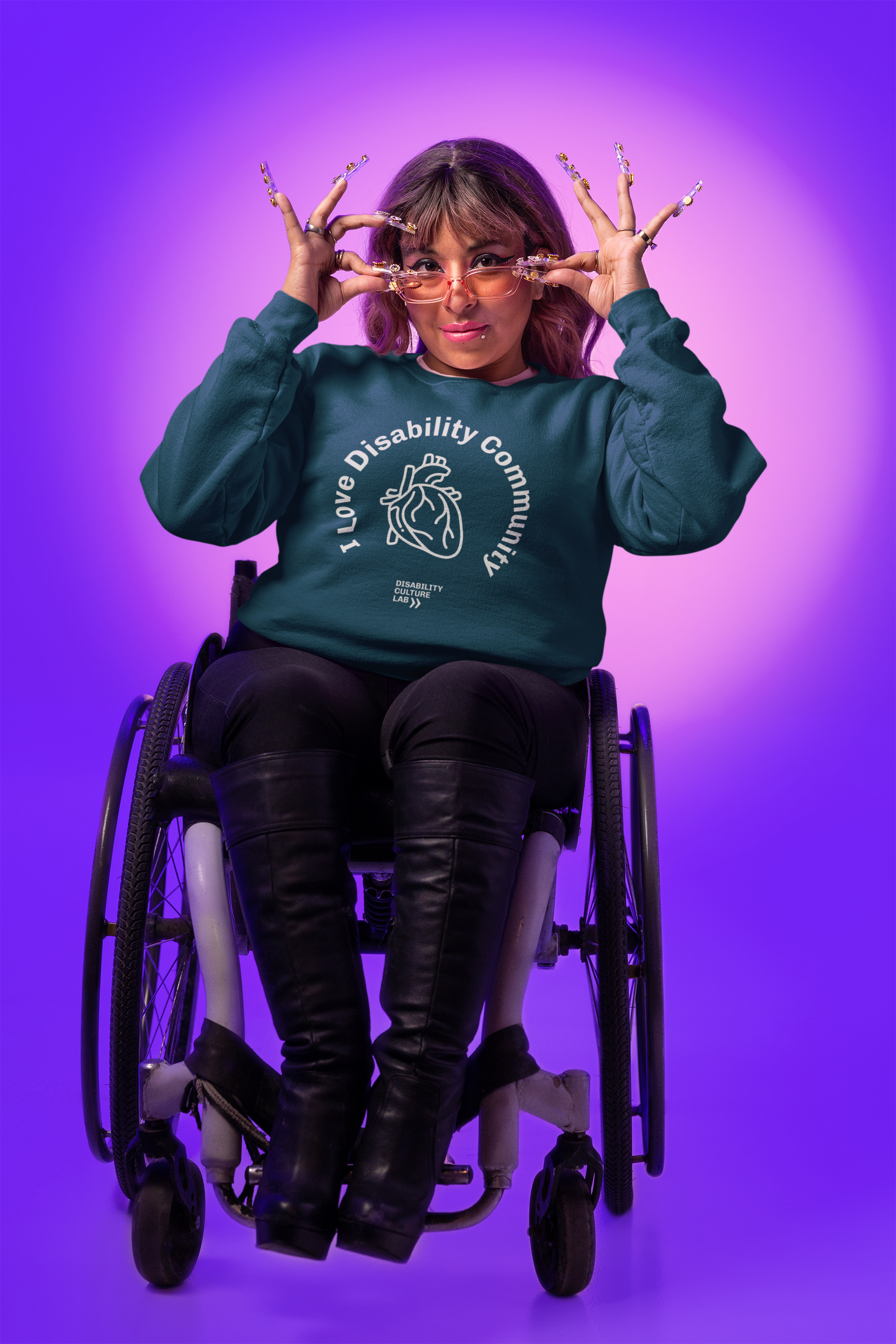 Against a purple backdrop, an individual in a wheelchair showcases the Disability Culture Lab's limited-edition collaboration, wearing the "I Heart Disability Community" sweatshirt in dark green. They complete the stylish look with chic glasses and elaborate long nail art, playfully gesturing near their face.