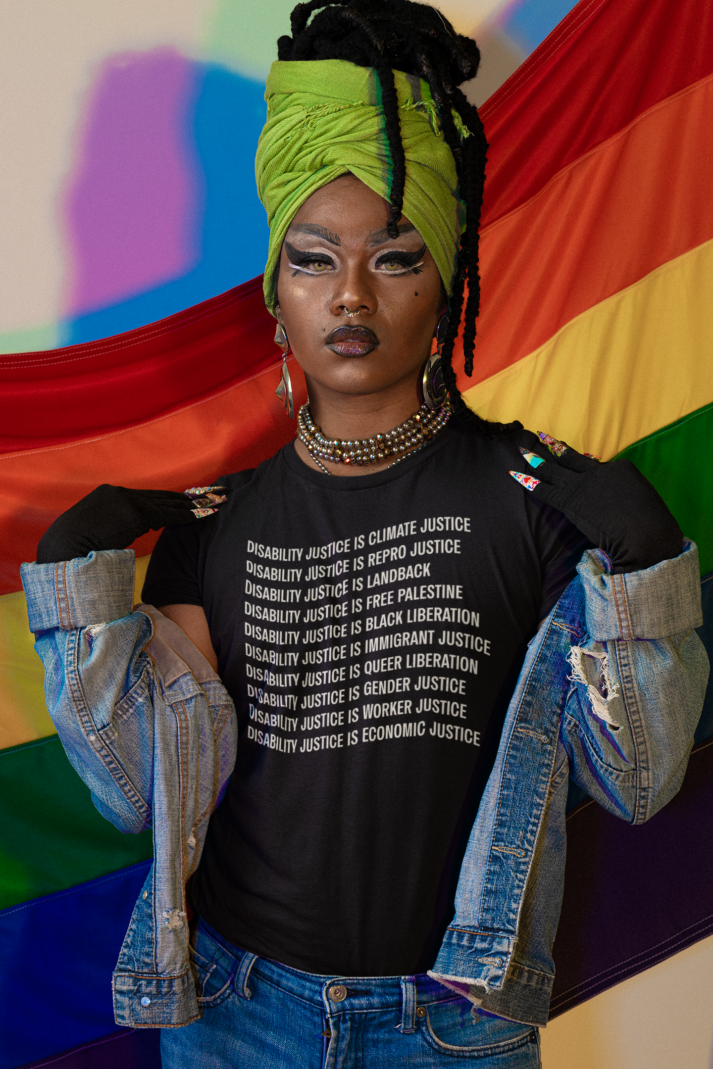 A person in a green headwrap and denim jacket poses confidently in front of a rainbow flag, wearing the "Disability Justice Is Tee" by Disability Culture Lab. The black T-shirt is emblazoned with powerful messages advocating for disability, climate, and gender justice, reflecting the bold spirit of Jennifer White-Johnson and the Disability Culture Lab.