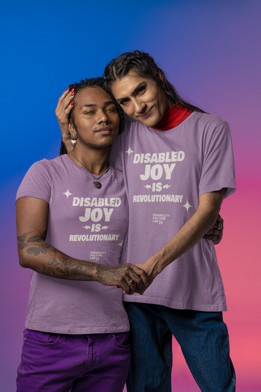 Two people stand together on a purple-to-pink gradient background, wearing matching limited-edition "Disability Joy Tee" shirts by Disability Culture Lab that read "Disabled joy is revolutionary." They hold hands as one gently rests their head on the other's shoulder, celebrating Disability Culture.