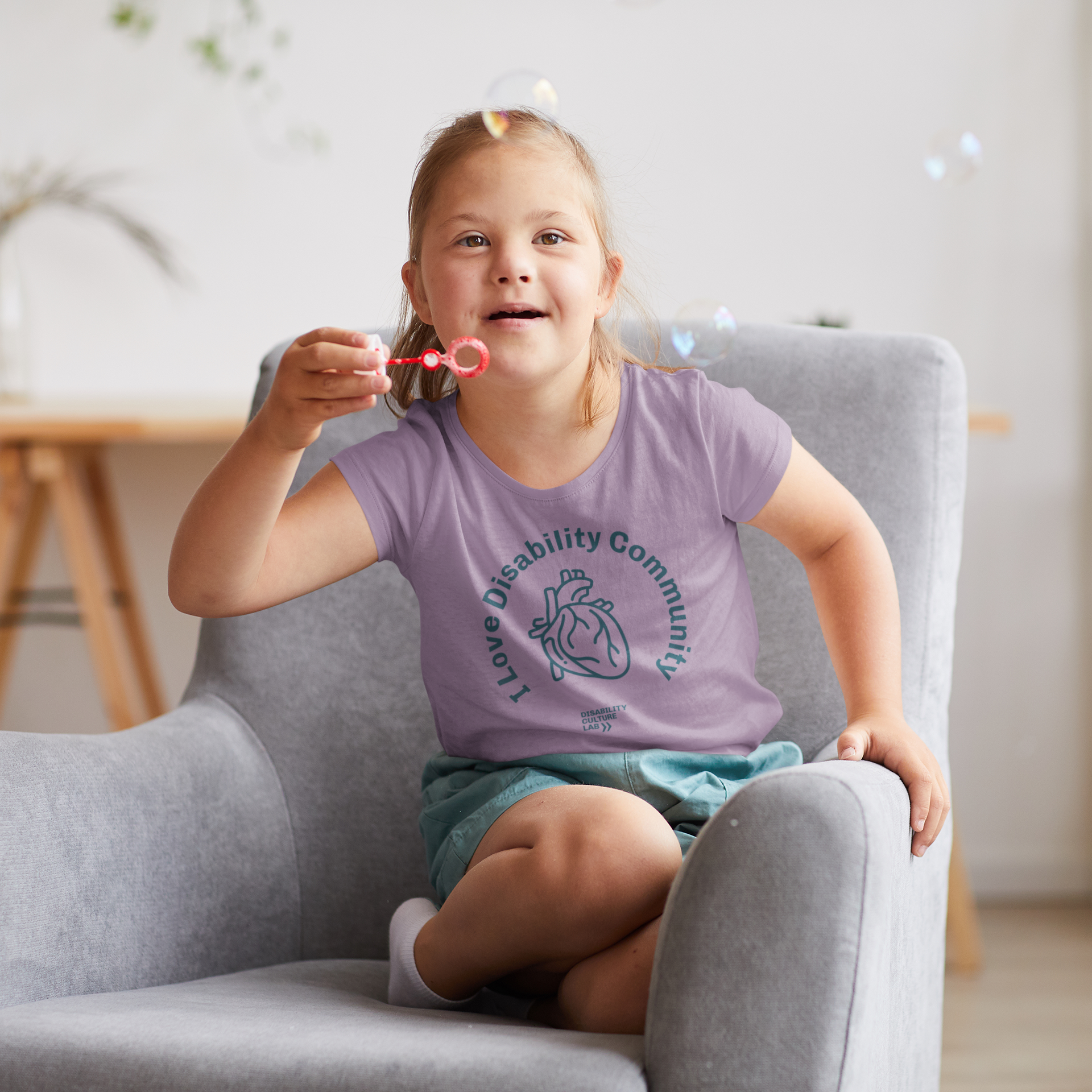 A young girl with Down syndrome sits joyfully in a gray armchair, blowing bubbles. She wears a lavender "I Heart Disability Community Kids Tee" from Disability Culture Lab, part of an exclusive collaboration to support the disability community. The softly lit room has a plant in the background.