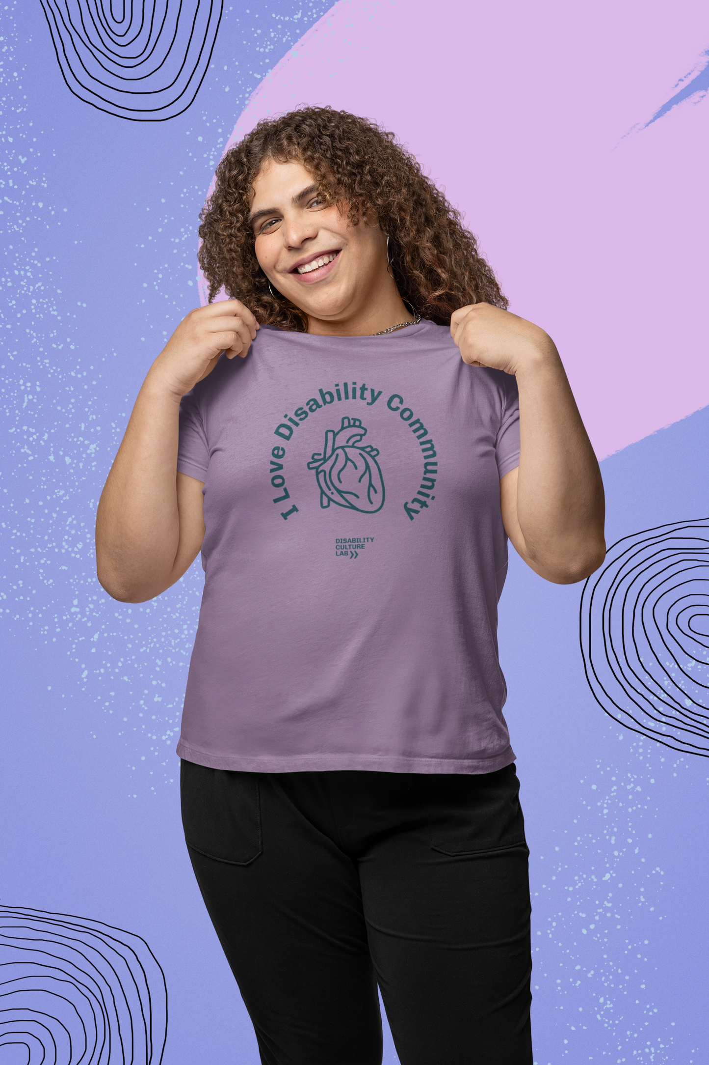 A person with curly hair smiles while grasping the collar of their purple I Heart Disability Community Tee by Disability Culture Lab. The T-shirt features a printed heart and the phrase "I love Disability Community" set against a backdrop of purple and blue abstract patterns.